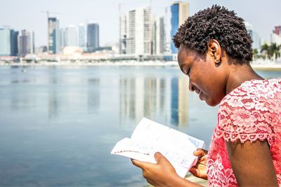 Side view of woman reading novel by sea on sunny day