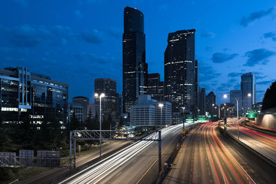 Light trails on road amidst buildings against sky at night