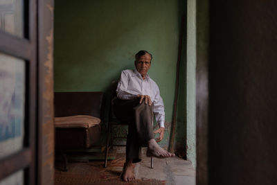 Portrait of man sitting on chair against wall at home seen through doorway