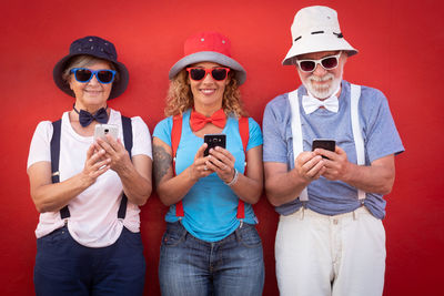 Smiling fashionable family using mobile phones while standing side by side against red wall