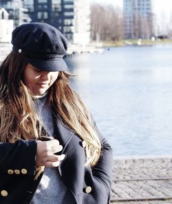 Woman in jacket and cap standing against lake in city