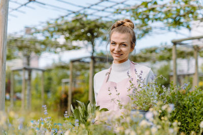 Farmer with a pot of fresh plants standing at her greenhouse and looking at the camera with smile