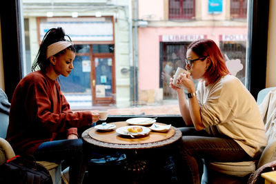 Side view of stylish enthusiastic multiethnic girl friends talking and drinking sitting at table nearby window in cafe