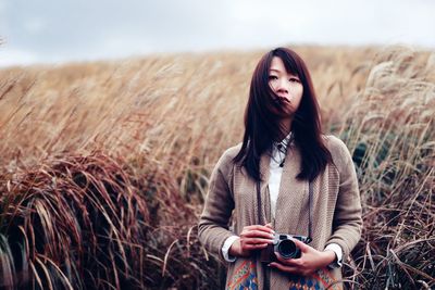 Portrait of young woman with camera standing on grassy field against sky