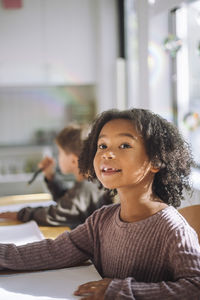 Smiling girl with curly hair looking away while sitting in classroom at kindergarten