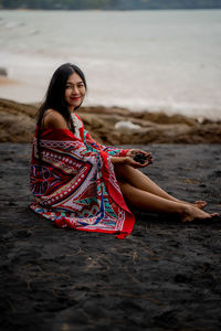 Portrait of smiling young woman with shawl sitting on black sand beach