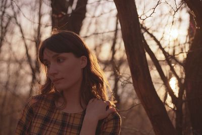 Woman looking away against bare trees during sunset