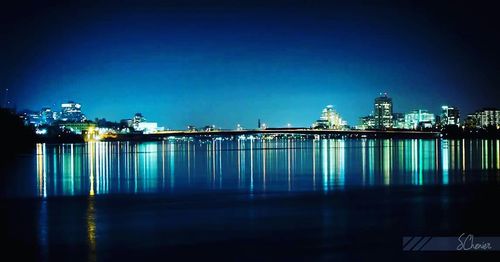 Illuminated buildings by river against clear blue sky at night