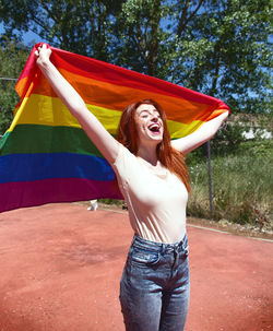 Cheerful young woman holding rainbow flag on basketball court against trees