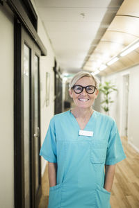 Portrait of mature female healthcare worker with hands in pockets standing in hospital corridor