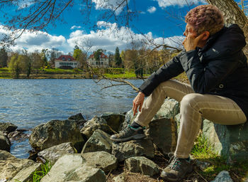 Man sitting on rock by river against sky