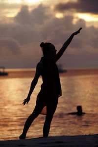 Silhouette woman with arms outstretched standing at beach against sky during sunset