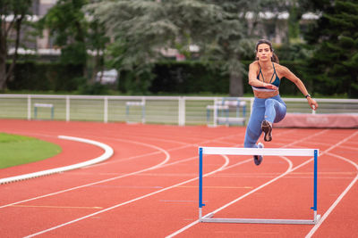 Full length of confident young hispanic sportswoman jumping over barrier while running on red track of sports stadium