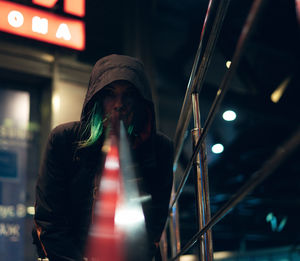 Low angle portrait of woman wearing hooded jacket standing by railing at night