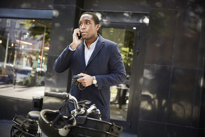 Young male commuter talking on smart phone while standing with electric bicycle against building in city