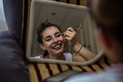 Reflection of cheerful young woman applying make-up on mirror at home