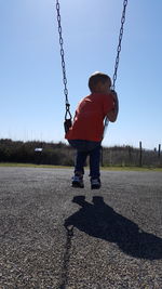 Low angle view of boy sitting on swing over road against sky during sunny day