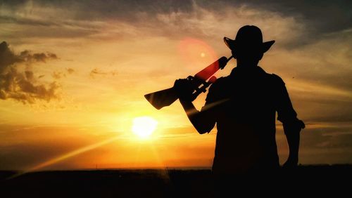 Silhouette man with rifle standing against orange sky