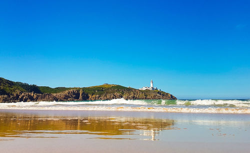 Lighthouse on beach by sea against clear blue sky
