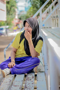 Young woman wearing hijab sitting on bench in city