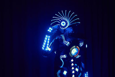 Faceless person in contemporary glowing suit of space cyborg with neon illumination and helmet standing on black background in dark studio person