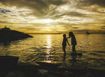 Silhouette sisters walking in sea against cloudy sky during sunset