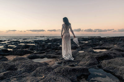 Woman standing on rock by sea against sky during sunset