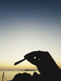 Silhouette person holding umbrella in front of sea against clear sky during sunset