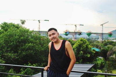 Smiling young man standing by railing against sky