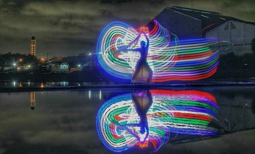 Digital composite image of illuminated light by river against buildings at night