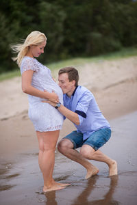 Man crouching while kissing stomach of pregnant wife at beach