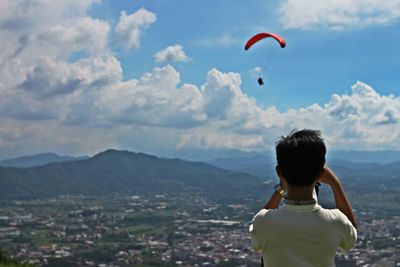 Rear view of man photographing paraglider flying over landscape against cloudy sky