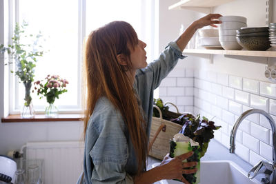 Side view of woman holding vegetables while looking at bowls on shelf in kitchen