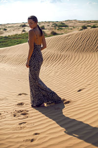 Brunette woman in a long leopard dress stands with her back in the desert at sunset. go everywhere