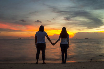 Rear view of couple standing at beach against sky during sunset