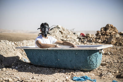 Portrait of young man wearing gas mask in bathtub against clear sky