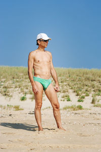 Shirtless mature man standing at beach against clear blue sky