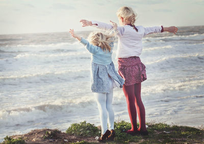 Rear view of sisters with arms outstretched standing on cliff against sea