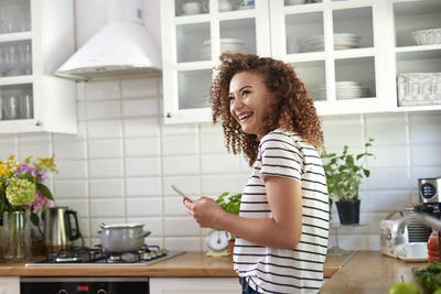 Side view of cheerful teenage girl using smart phone while standing in kitchen at home