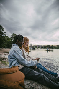 Side view of teenage boy using smart phone with female friend while sitting on rock under cloudy sky