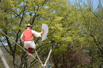 Low angle view of tightrope walker holding hand fan against trees