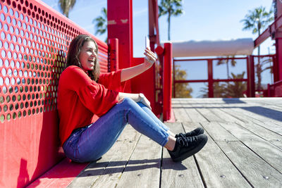 Teenage girl taking selfie with mobile phone while sitting against railing during sunny day