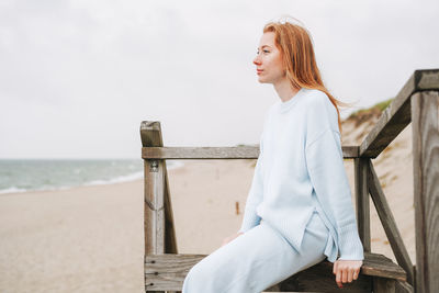 Portrait of young red haired woman in light blue sweater on sand beach by sea in storm