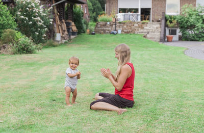 Full length of mother sitting on grass while looking at cheerful son in yard