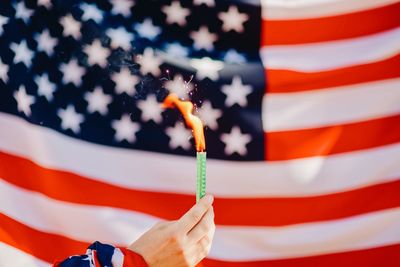 Cropped hand of person holding illuminated firework against american flag