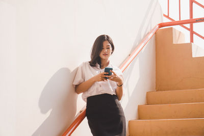 Young woman using mobile phone while standing against wall