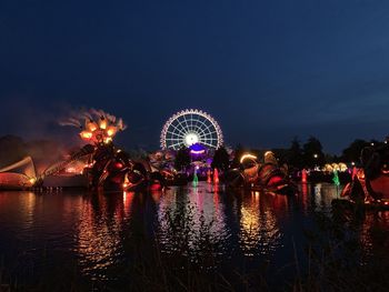 Illuminated ferris wheel by river against sky at night