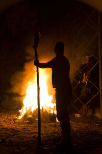 Silhouette man holding bamboo while looking at fire during night