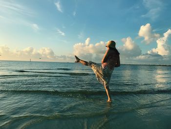 Rear view of woman with leg up standing on shore at beach against sky