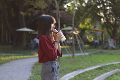 Side view of woman drinking coffee while standing on field against trees in park